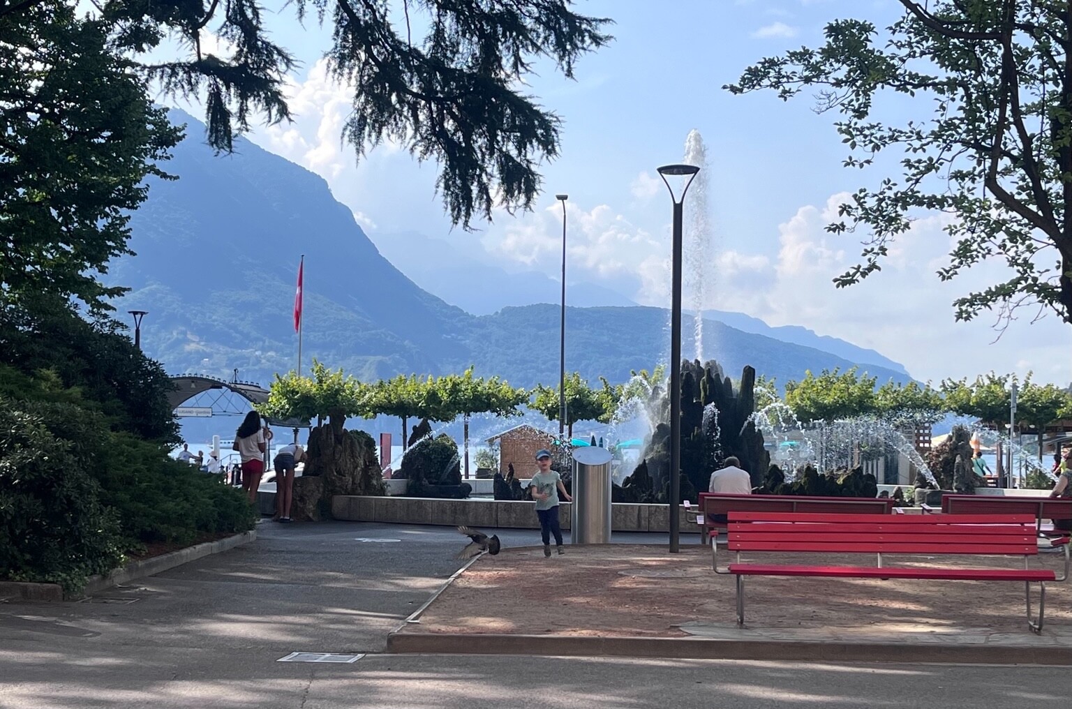 View on mountains over the lake, blue sly with clouds coming over the mountains. Foreground red bench and a rocky fountain just before green roof-like trees.